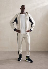 Sudadera crema con acentos azul oscuro, combinada con pantalones jogger crema a juego y zapatillas negras con detalles en amarillo neón. Textura suave.