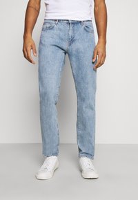 Homem com jeans de denim azul claro, ténis brancos e uma camisa branca, de pé contra um fundo simples cinza claro.