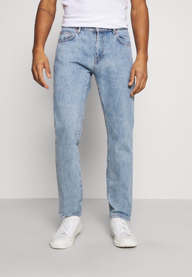 Homem com jeans de denim azul claro, ténis brancos e uma camisa branca, de pé contra um fundo simples cinza claro.