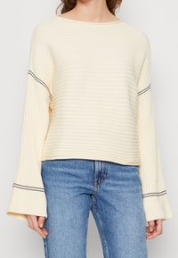 Cream ribbed sweater with wide bell sleeves, contrasting black stitching along the sleeve cuffs, paired with blue denim jeans.