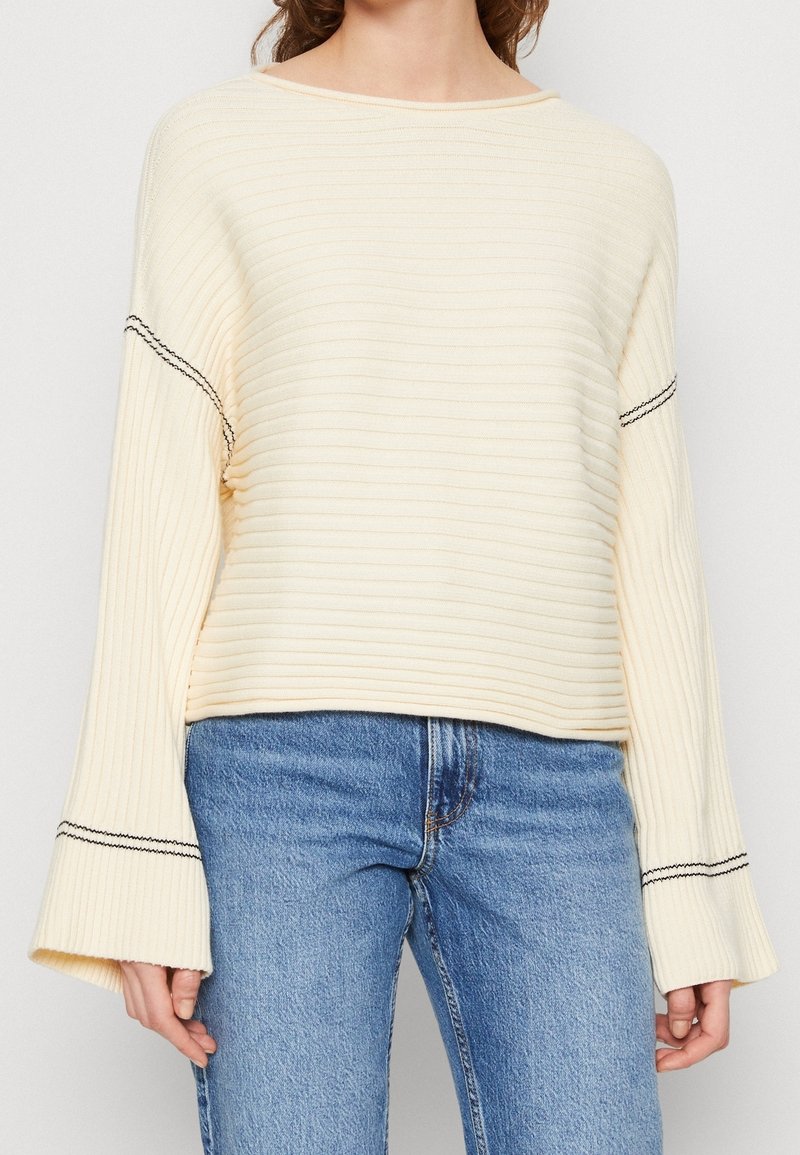 Cream ribbed sweater with wide bell sleeves, contrasting black stitching along the sleeve cuffs, paired with blue denim jeans.