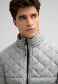 Giacca trapuntata grigia chiara con colletto alto, dotata di zip frontale e accessori neri; texture morbida e motivo a diamante su tutta la superficie.