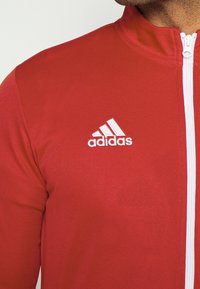 Homme portant une veste de sport zippée rouge Adidas avec un logo blanc sur la poitrine gauche et des détails de fermeture éclair blancs.