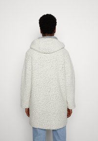 Abrigo blanco y esponjoso con un material texturizado similar al borrego. Presenta una gran capucha y mangas anchas, con costuras visibles a lo largo de los bordes.