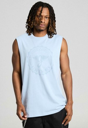 Man with dreadlocks wearing a sleeveless light blue shirt with a faint circular graphic and black pants against a plain background.