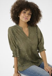 Olīvu zaļa mežģīņu blūze ar ziedu rakstu, V veida kakla izgriezumu un īsām puff piedurknēm. Vieglas auduma ar sarežģītām izgriezumu detaļām un maigu tekstūru.