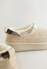 Beige suède instapper met faux fur voering, voorzien van een gedessineerde accentrand en decoratieve stiksels. Platte rubberen zool.