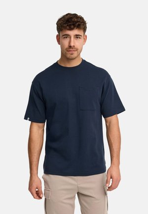 Mann mit kurzen, lockigen braunen Haaren trägt ein marineblaues Kurzarmhemd und beige Cargo-Hosen, steht vor einem weißen Hintergrund.