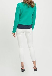 Zielona pikowana kurtka noszona na granatowej bluzce, w parze z białymi obcisłymi dżinsami i czarnymi sandałami na wysokim obcasie, widok z tyłu.