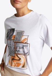 Camiseta de algodón blanca que presenta un collage de impresiones gráficas en negro, gris y tonos tierra, con costuras finas y un ajuste relajado.