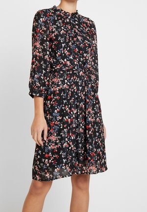 Kobieta ubrana w czarną sukienkę do kolan z czerwono-niebiesko-szarym kwiatowym wzorem i rękawami długości za łokieć, zawiązanym paskiem w talii, stojąca na jednolitym tle.