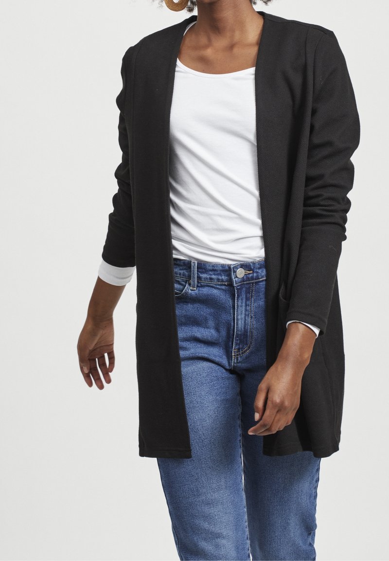 Femme portant un cardigan noir ouvert, une chemise blanche et un jean bleu, debout devant un fond clair uni.