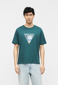 Young man wearing teal Jack & Jones T-shirt with beach graphic and light blue jeans, standing against plain light background.