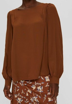 Donna che indossa una blusa marrone a maniche lunghe con maniche a sbuffo e una gonna marrone con motivo floreale con fiori bianchi e blu.