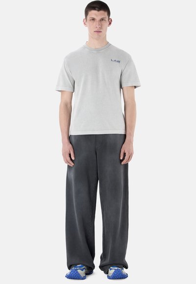 Jeune homme debout portant une chemise à manches courtes gris clair avec le logo "LAB", un pantalon ample gris foncé et des chaussures de sport bleues texturées sur fond blanc.