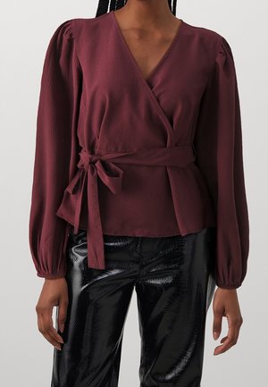 Blouse portefeuille bordeaux avec manches longues bouffantes, tissu texturé et taille nouée ; associée à un pantalon noir brillant à motifs.