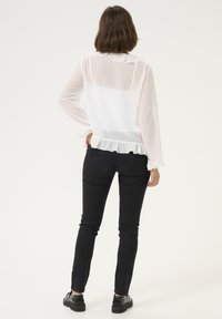 Hvid gennemsigtig bluse med flæser ved kraven og kanten, parret med sort tætsiddende jeans og sorte sko. Blusen har lange ærmer og en struktureret overflade.