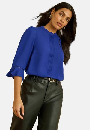 Königsblaue Bluse mit geschwungenem Ausschnitt, strukturierten vertikalen Falten und dreiviertellangen Puffärmeln; kombiniert mit hoch taillierter schwarzer Kunstlederhose.