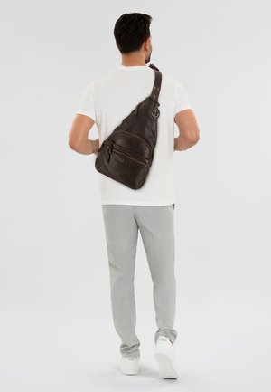 Borsa a tracolla in pelle marrone con finitura texture, dotata di tasca con zip, tracolla regolabile e un emblema ad ancora, indossata sulla schiena.