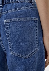 Jeans de mezclilla con un clásico bolsillo trasero con una costura reforzada, de color azul medio y una textura ligeramente desgastada.