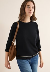 Haut noir à manches 3/4 avec des accents blancs, fabriqué dans un matériau doux. Associé à un jean en denim et un sac bandoulière marron. Tenue décontractée.