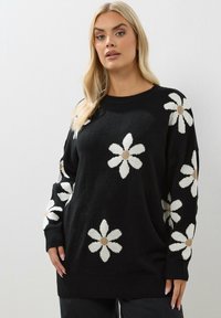 Schwarzer Strickpullover mit großen weißen Gänseblümchen-Mustern und beigen Mittelpunkten; lockere Passform mit gerippten Bündchen und Saum.