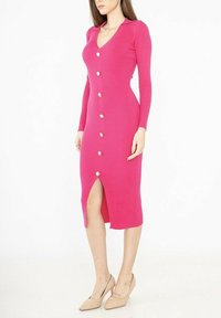 Robe longue en tricot côtelé rose avec un décolleté en V, des boutons sur le devant et une fente sur le côté. La robe est dotée d'un col et est associée à des talons beiges.