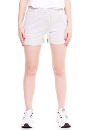 Une personne pose avec un short blanc à cordon de serrage et des baskets blanches sur un fond blanc uni.