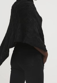 Personne portant un châle en tricot noir texturé drapé sur les épaules et un jean noir ajusté, debout devant un fond clair uni.