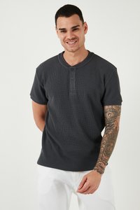 Hombre sonriente con camisa de manga corta abotonada de color gris oscuro y pantalones blancos, mostrando un tatuaje detallado que cubre todo el brazo derecho.