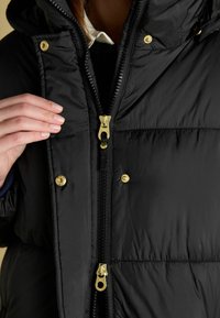 Černá péřová bunda s rozepínáním na zip a zlatými detaily, s kapucí, nadýchanou strukturou a patentkami po stranách zipu.