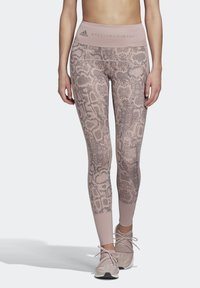 Žena v růžových legínách s vysokým pasem se vzorem hadí kůže a světle růžových sportovních botách stojí před jednobarevným pozadím.