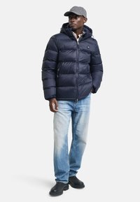 Marineblå dunjakke med hette, quiltet design, to glidelåslommer, brukt over en rutete skjorte, kombinert med lyseblå jeans og svarte støvler.