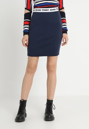 Jupe ajustée bleu marine en tissu doux, avec une taille haute arborant un logo "TOMMY JEANS". Portée avec un haut à rayures à manches longues et des bottes noires.