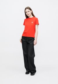 Κόκκινη βαμβακερή μπλούζα με στρογγυλή λαιμόκοψη και λογότυπο Calvin Klein. Συνδυάζεται με σκούρο τζιν παντελόνι φαρδιάς γραμμής και μαύρα παπούτσια. Απλό φόντο.