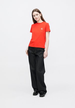 Červené bavlnené tričko s okrúhlym výstrihom a logom Calvin Klein. Kombinované s tmavými širokými džínsovými nohavicami a čiernymi topánkami. Jednofarebné pozadie.