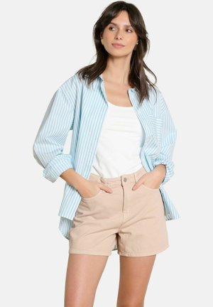 Mujer con pantalones cortos beige, camiseta sin mangas blanca y camisa suelta a rayas azules y blancas, con las manos en los bolsillos, mirando ligeramente hacia un lado.