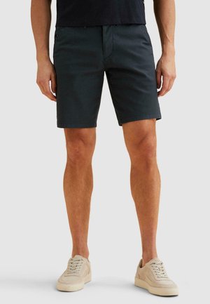 Mann trägt schwarze knielange Shorts, schwarzes Hemd und beige Freizeitschuhe, steht vor einem schlichten hellgrauen Hintergrund.