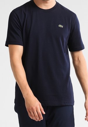 Mann trägt ein marineblaues kurzärmeliges T-Shirt mit einem kleinen grünen Krokodil-Logo auf der linken Brust.