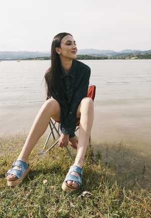 Donna con capelli lunghi e scuri, indossa una giacca scura e sandali di jeans, seduta su una sedia rossa vicino a un lago con montagne sullo sfondo.