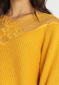 Żółty sweter z dzianiny z koronkową nakładką przy dekolcie, z teksturowanym wzorem i luźnym krojem. Koronka dodaje delikatny detal.