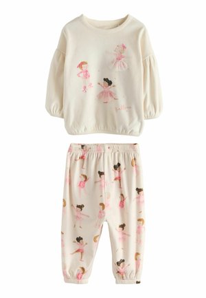 Lille barn med krøllet hår iført cremefarvet pyjamas med ballerina-design, stående barfodet på en hvid seng i et lyst soveværelse.