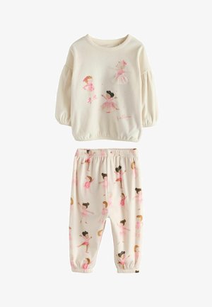 Lille barn med krøllet hår iført cremefarvet pyjamas med ballerina-design, stående barfodet på en hvid seng i et lyst soveværelse.