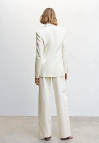 Personne aux cheveux blonds courts portant un costume entièrement blanc avec un pantalon à jambes larges et des talons lavande, debout contre un mur clair uni.