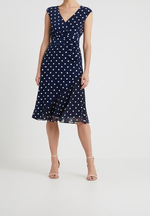 Femme portant une robe portefeuille bleu marine sans manches à pois blancs et des talons beige à bride cheville ouverte, debout sur un sol blanc.