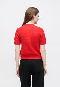 Czerwony sweter z krótkim rękawem i okrągłym dekoltem, wykonany z miękkiego, teksturowanego materiału. Noszony z wysokim stanem czarnych spodni. Bez widocznych wzorów ani detali.