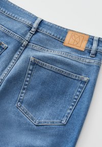 Jeans en denim bleu avec une pièce en cuir beige au niveau de la taille à l'arrière, deux poches arrière et une finition lisse et texturée.