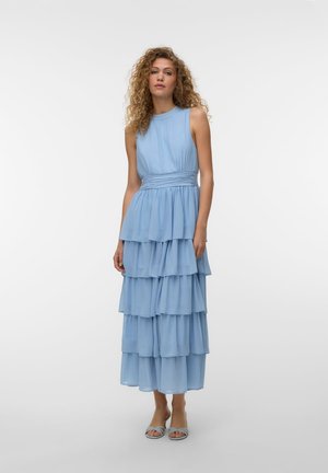Femme aux cheveux bouclés portant une robe à volants bleu clair sans manches et des sandales ouvertes assorties, debout devant un fond blanc uni.