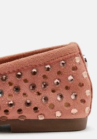 Zapato de ante rosa de tipo slip-on con forma redondeada, que cuenta con tachuelas doradas y en tono rosa a lo largo de la superficie para añadir textura. Suela plana marrón.