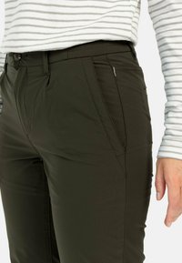 Dunkelgrüne Hose mit schmaler Passform, ausgestattet mit Taschen vorne und subtilen Stichdetails. Getragen mit einem hell gestreiften Langarmhemd.